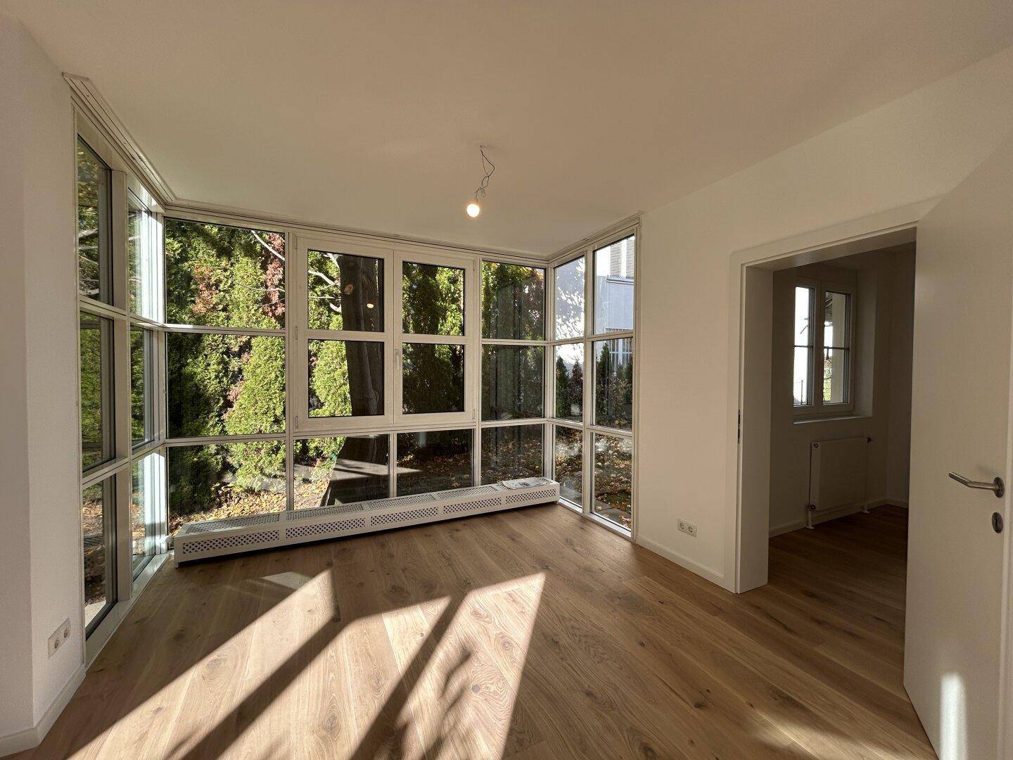 Heller Raum mit großen Fenstern, Holzboden und Blick ins Grüne.