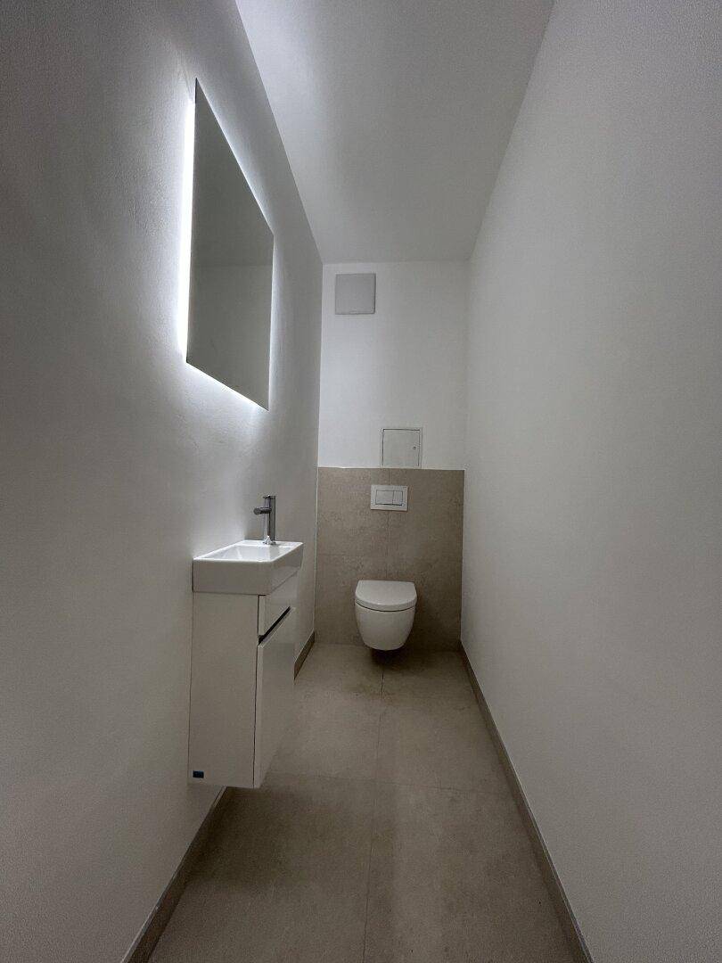Schmale Gästetoilette mit wandhängendem WC, kleinem Waschbecken und beleuchtetem Spiegel.