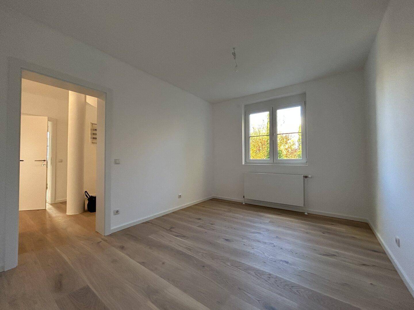Heller, leerer Raum mit Holzboden, Fenster und Blick ins Grüne.