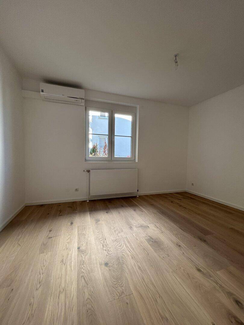 Heller, leerer Raum mit Holzboden, Fenster und Klimaanlage.