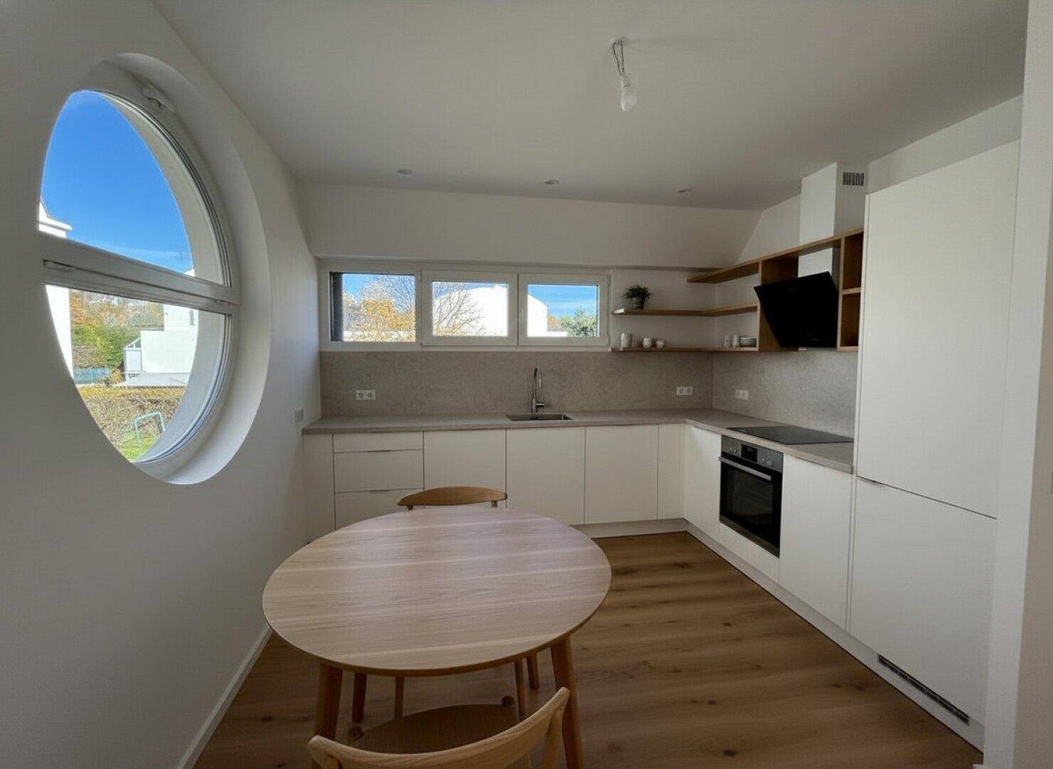 Moderne Küche mit weißen Schränken, Holzboden und einem runden Fenster, ideal für eine Essecke.