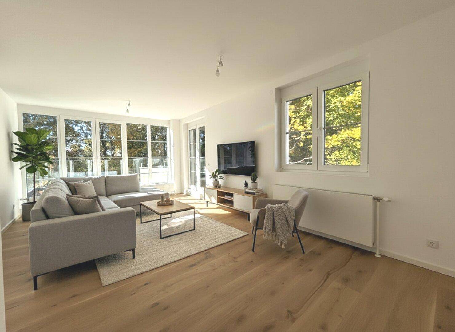 Helles Wohnzimmer mit Holzboden, modernem Sofa und großen Fenstern mit Blick ins Grüne.