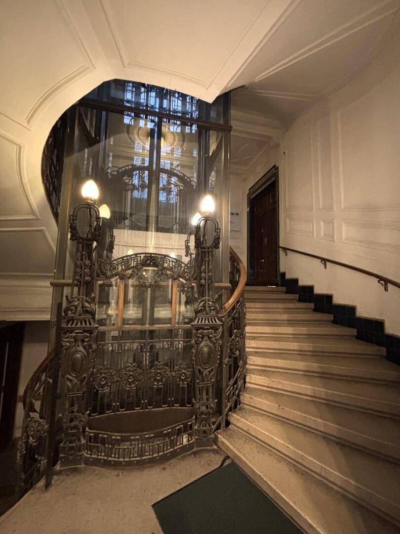 Elegantes Treppenhaus mit historischem Aufzug, kunstvollem Geländer und Marmorstufen.