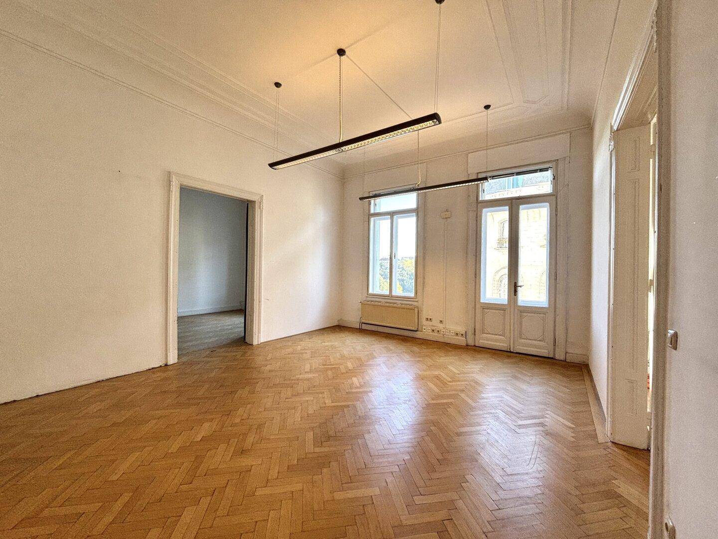 Heller, leerer Büroraum mit Fischgrätparkett, hohen Fenstern und klassischer Deckenverzierung.