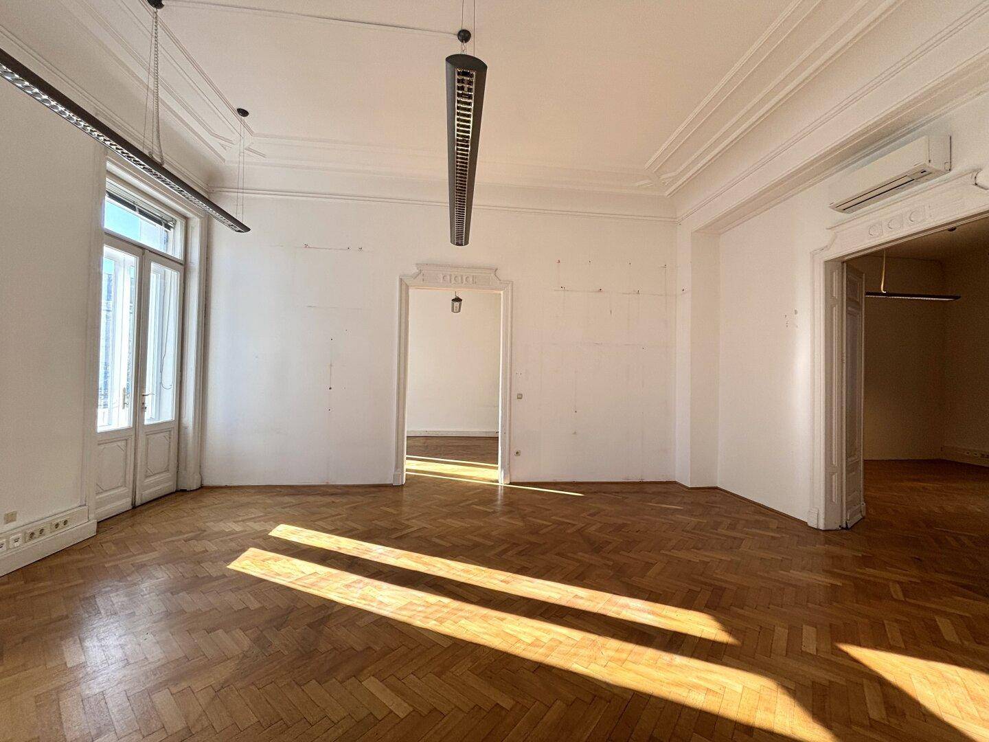 Großer, leerer Büroraum mit Fischgrätparkett, hohen Decken und Sonnenlicht, das durch die Fenster fällt.