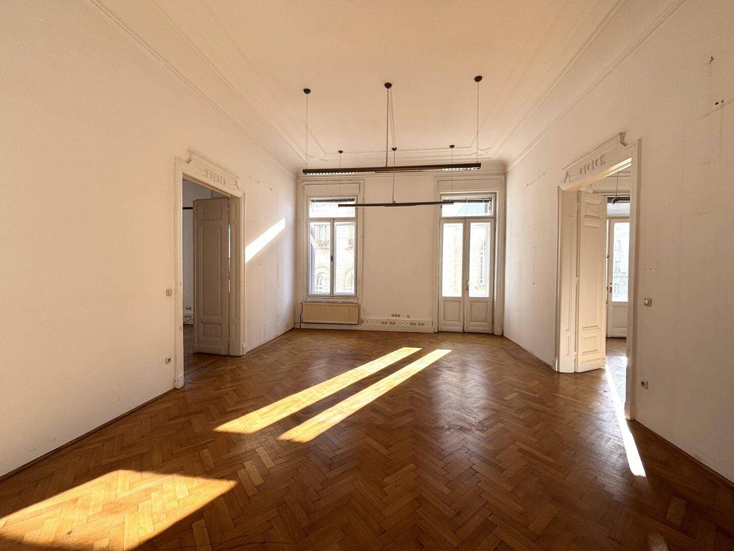 Heller, leerer Büroraum mit Fischgrätparkett, hohen Fenstern und Sonnenlicht, das durch die Fenster fällt.