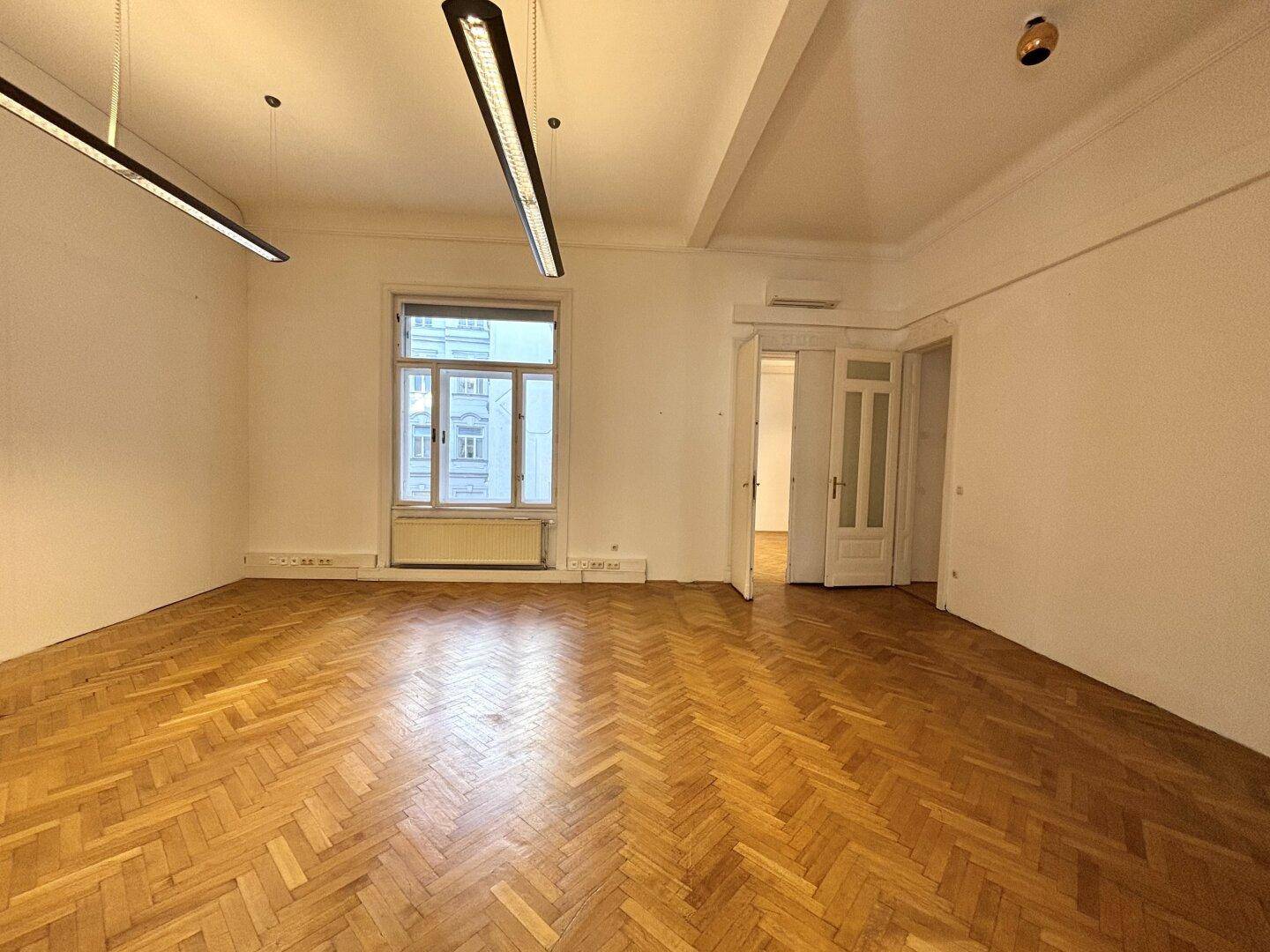 Leerer Büroraum mit Fischgrätparkett, hohen Decken und einem großen Fenster mit Vorhängen.