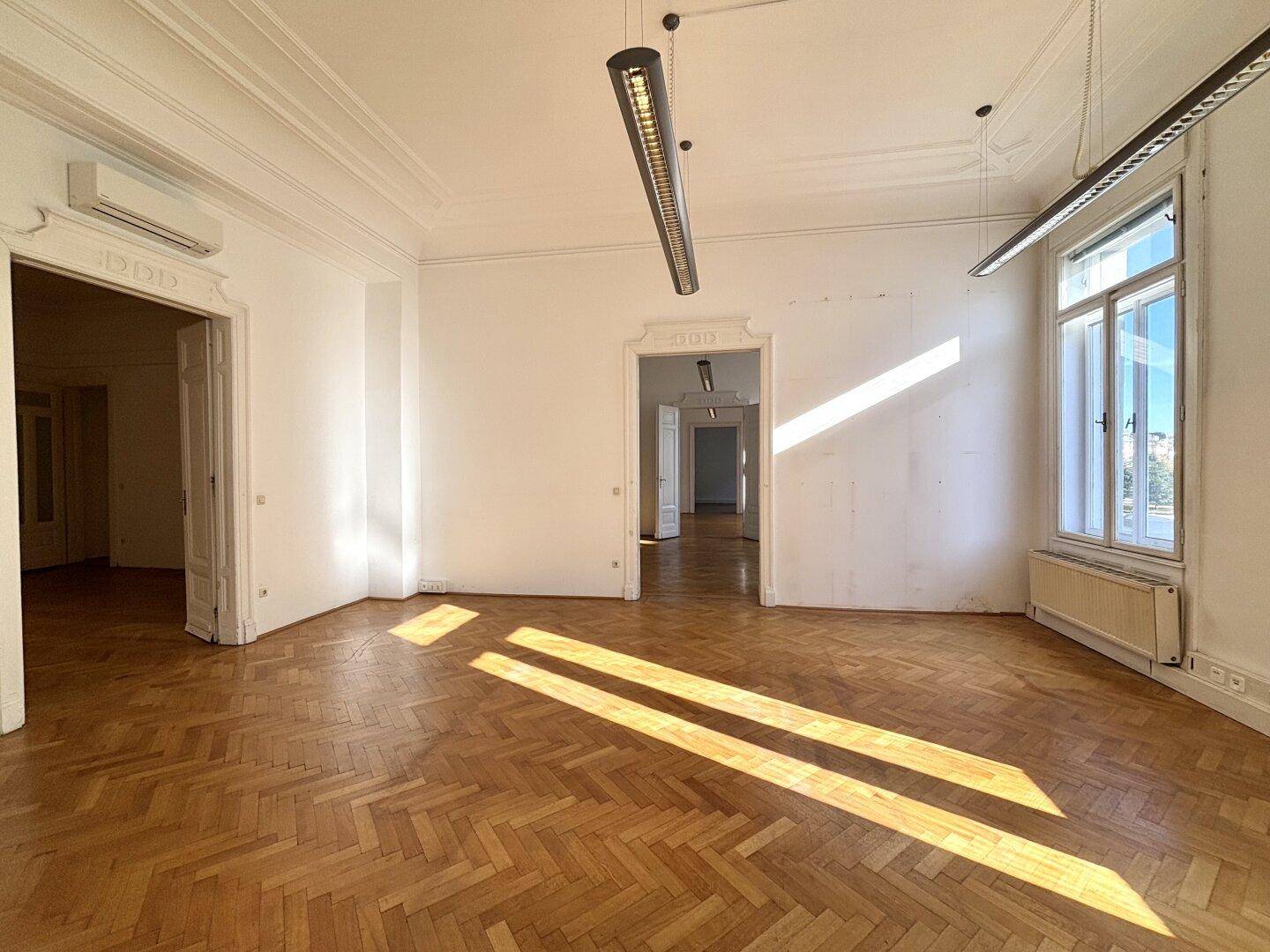 Großer, leerer Büroraum mit Fischgrätparkett, hohen Decken und Sonnenlicht, das durch die Fenster fällt.