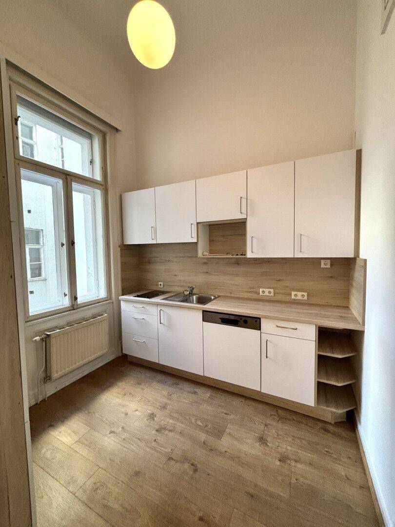 Kleine, helle Küche mit weißen Schränken, Holzoptik-Arbeitsplatte und Fenster mit Heizkörper.