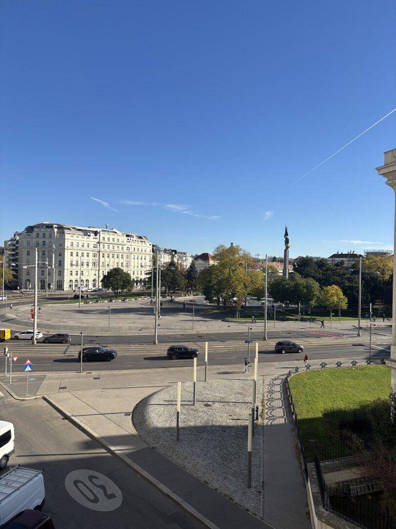 Blick von oben auf eine belebte Stadtkreuzung mit Gebäuden und Bäumen unter blauem Himmel.