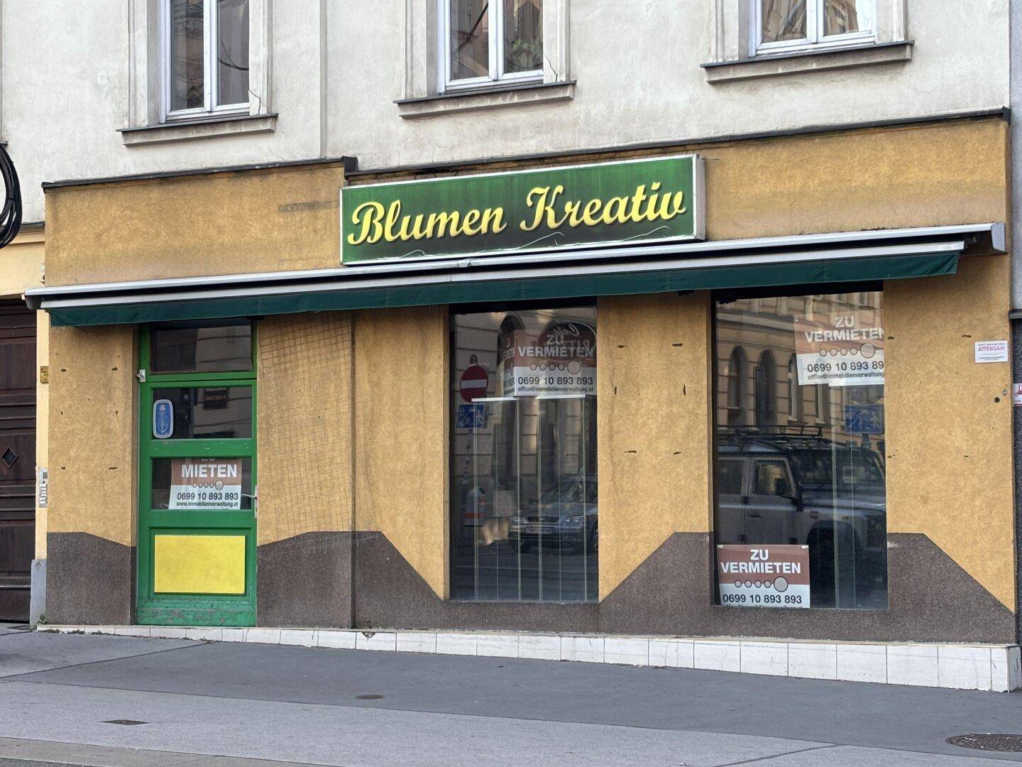 Außenansicht eines Geschäftsgebäudes mit Schaufenstern, einer Markise und einem Schild für "Blumen Kreativ".