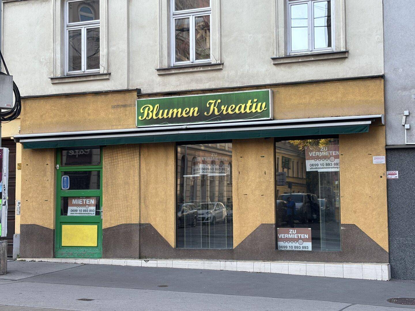 Außenansicht eines Geschäftsgebäudes mit Schaufenstern, einer Markise und einem Schild für "Blumen Kreativ".