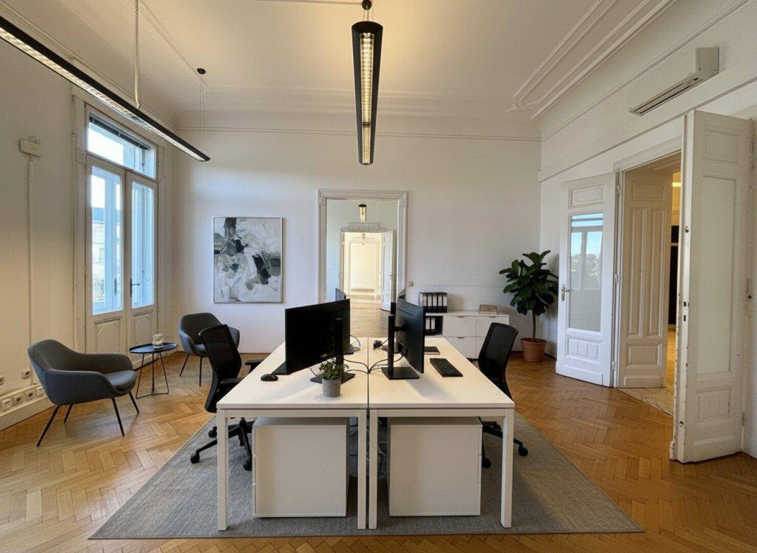 Modernes Büro mit zwei Arbeitsplätzen, Parkettboden und hohen Fenstern, die viel Licht hereinlassen.