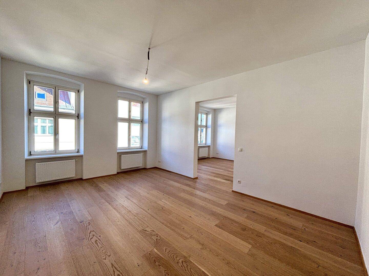 Geräumiger, heller Raum mit Holzboden und Fenstern, der in einen weiteren Bereich übergeht.