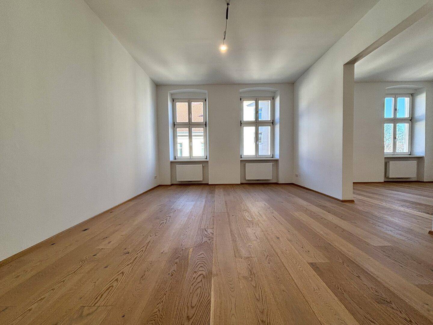 Leerer, heller Raum mit Holzboden und zwei großen Fenstern, bereit zur individuellen Gestaltung.