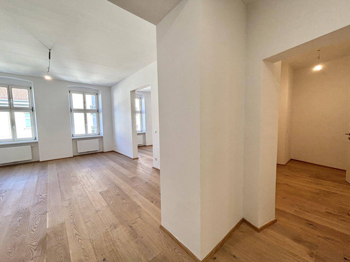 Heller Raum mit Holzboden und Fenstern, der offen in einen weiteren Bereich übergeht.