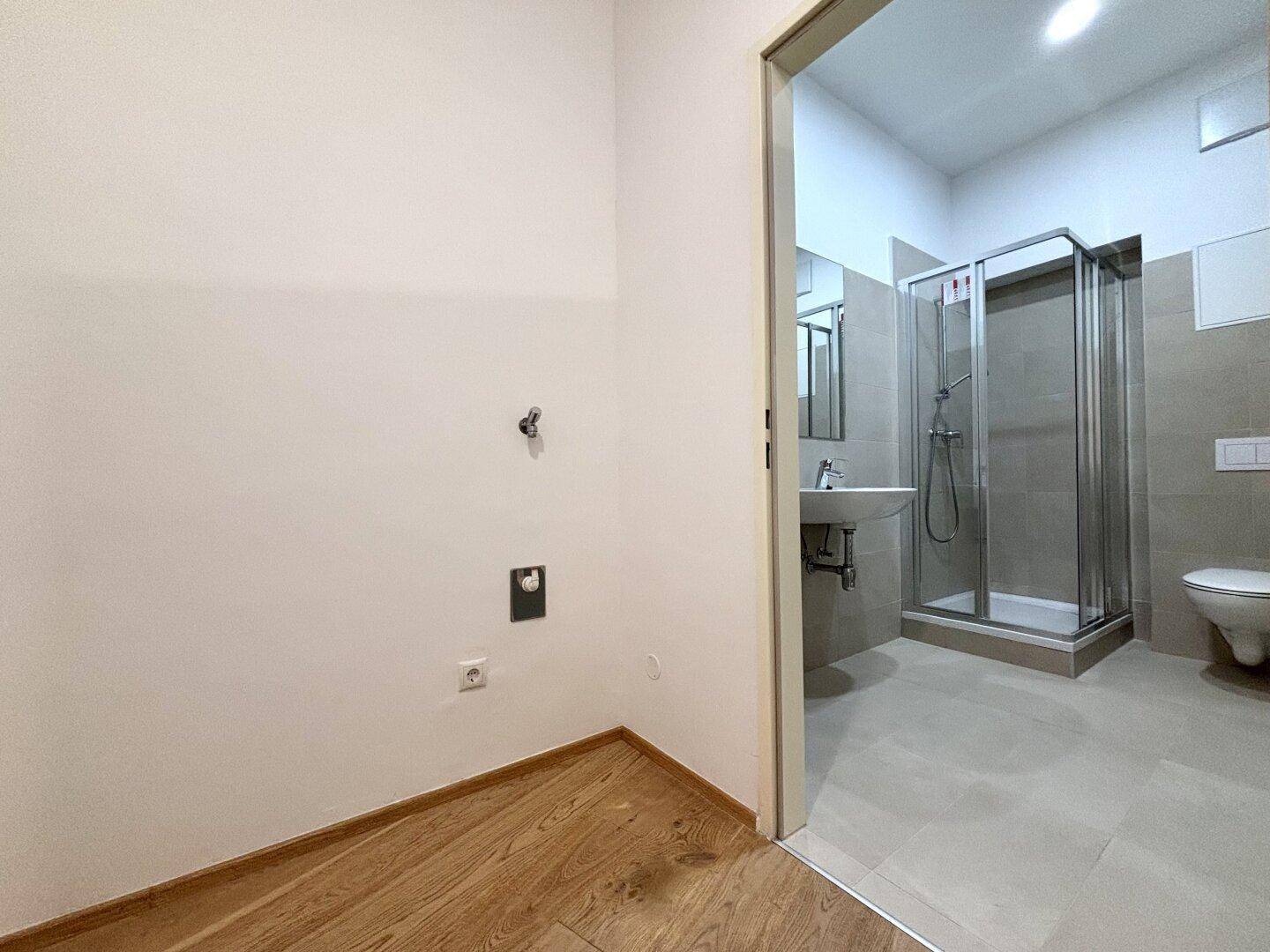 Blick von einem Raum mit Holzboden in ein modernes Badezimmer mit Dusche und Fliesen.