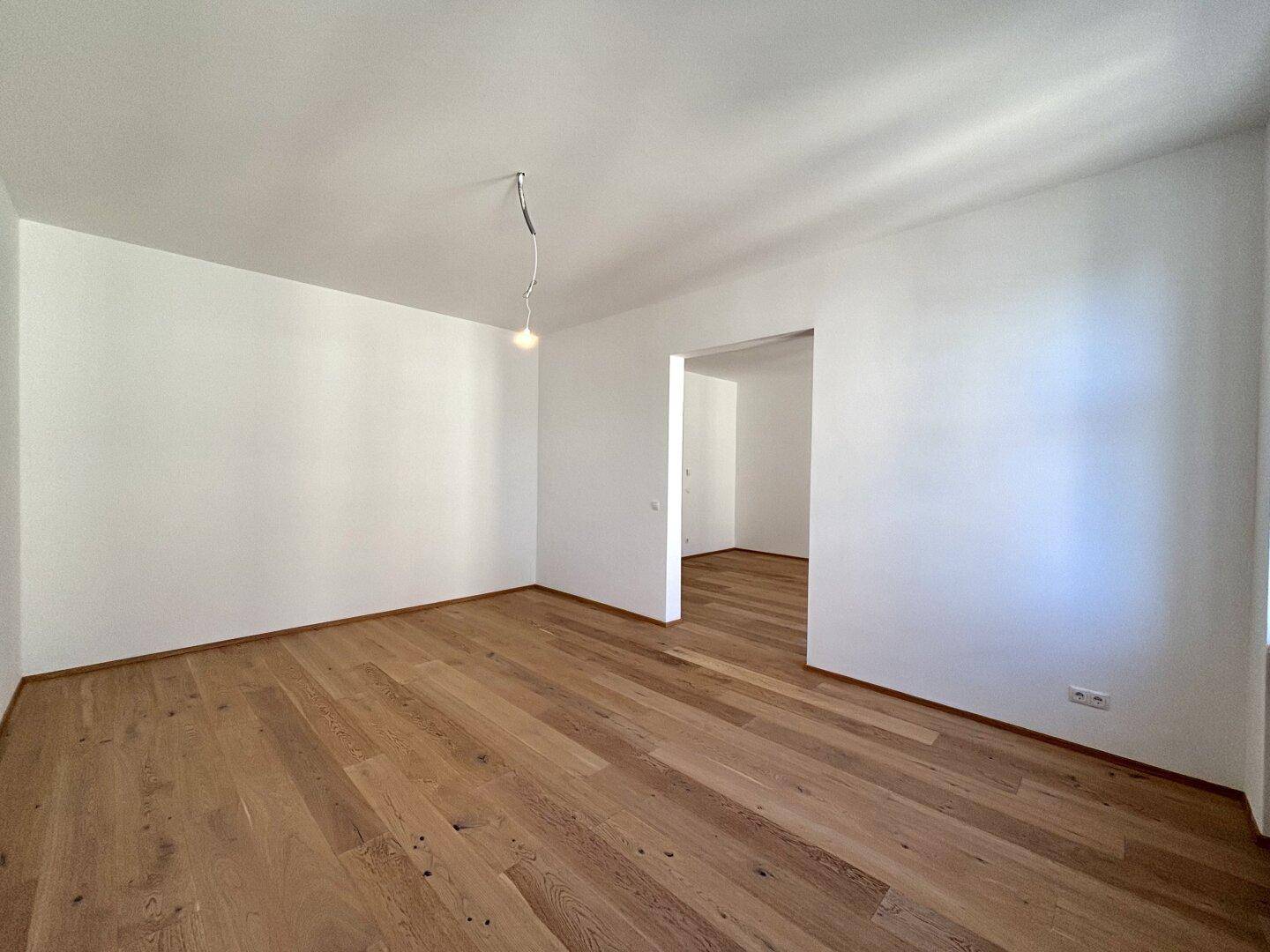 Heller, leerer Raum mit Holzboden und einem Durchgang zu einem weiteren Zimmer.