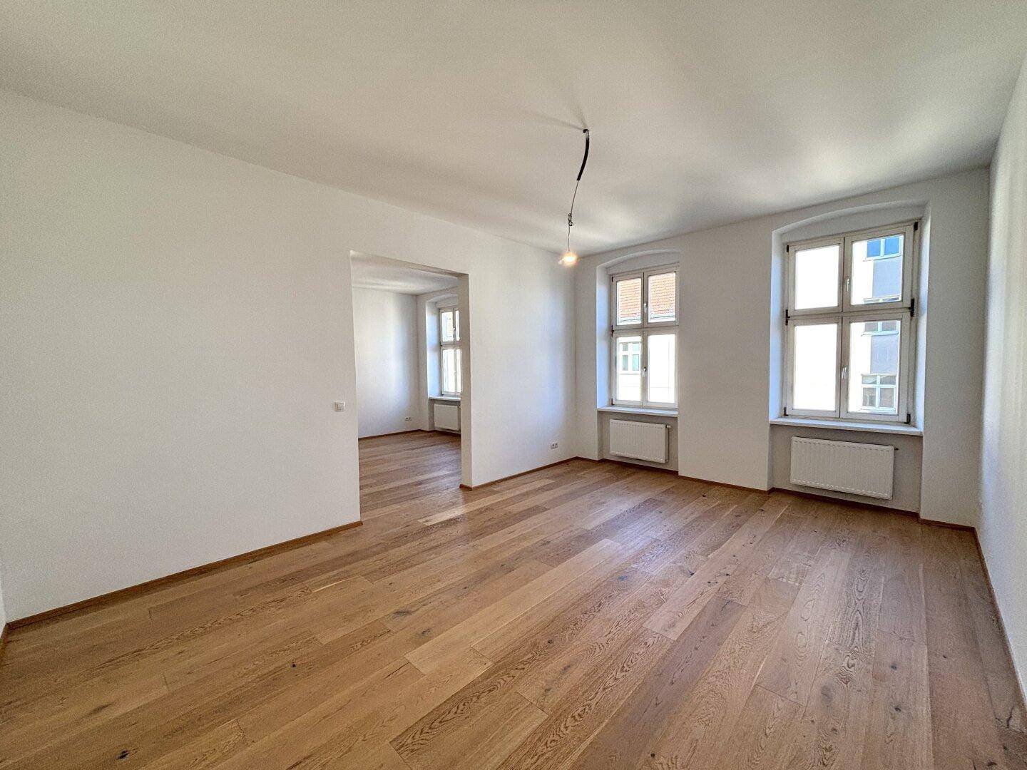 Heller Raum mit Holzboden und Fenstern, die einen Blick in einen weiteren Raum ermöglichen.