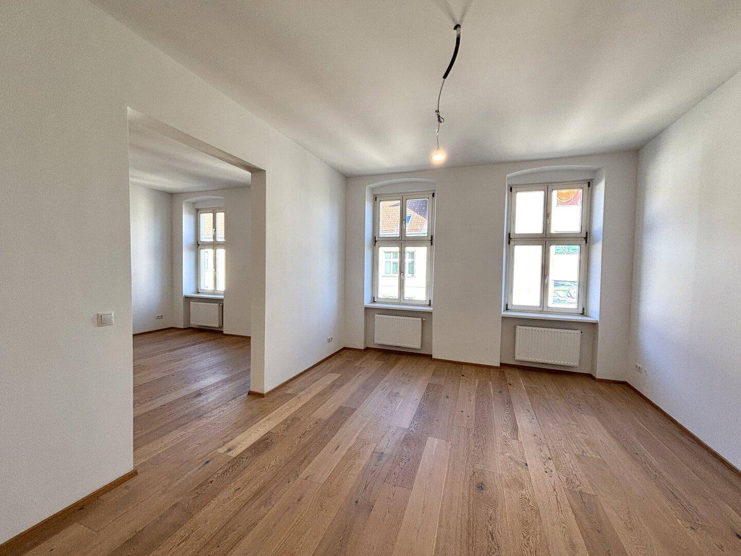 Heller Raum mit Holzboden und zwei Fenstern, der offen in einen weiteren Bereich übergeht.