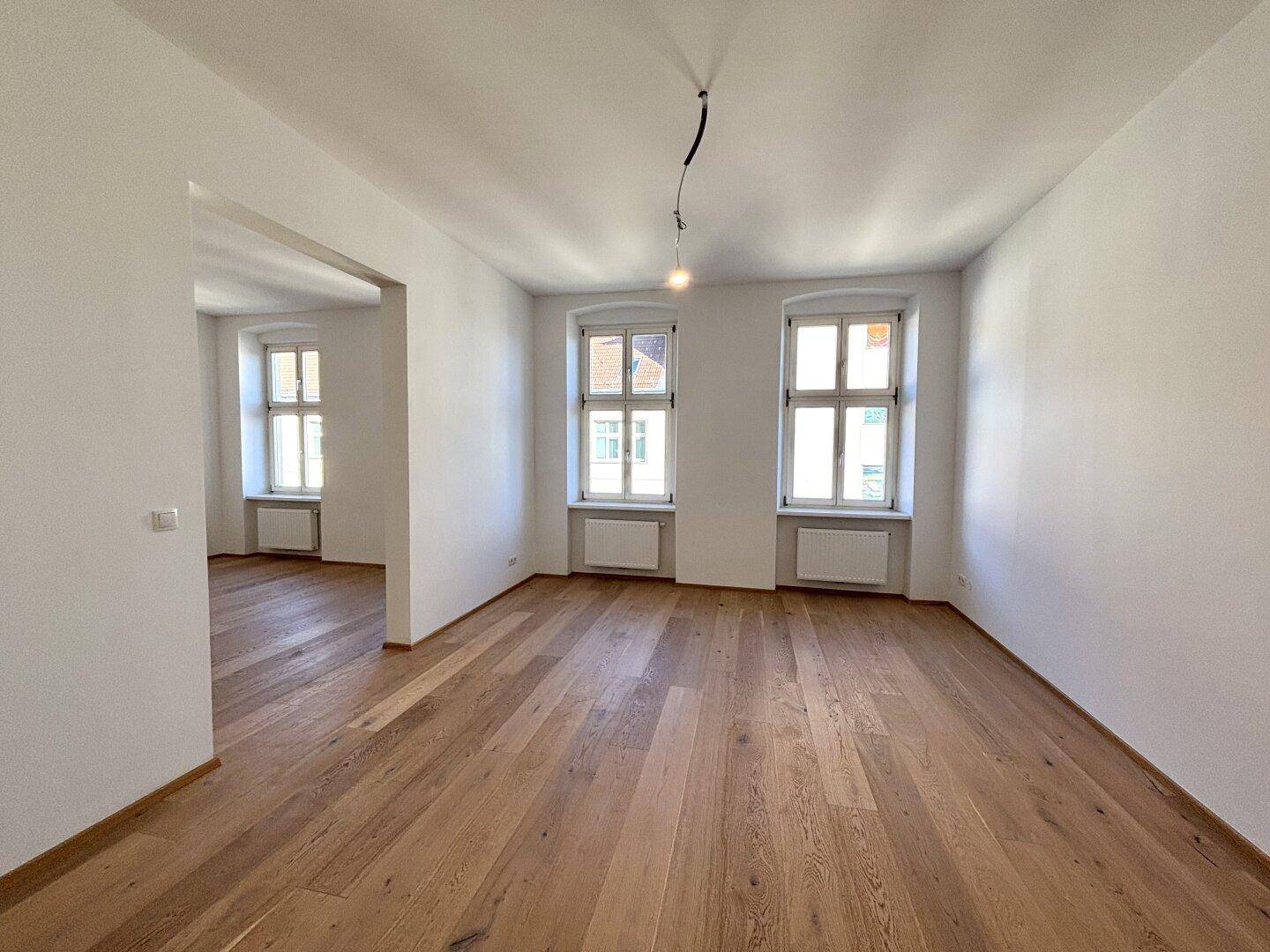 Heller Raum mit Holzboden und zwei Fenstern, der offen in einen weiteren Bereich übergeht.