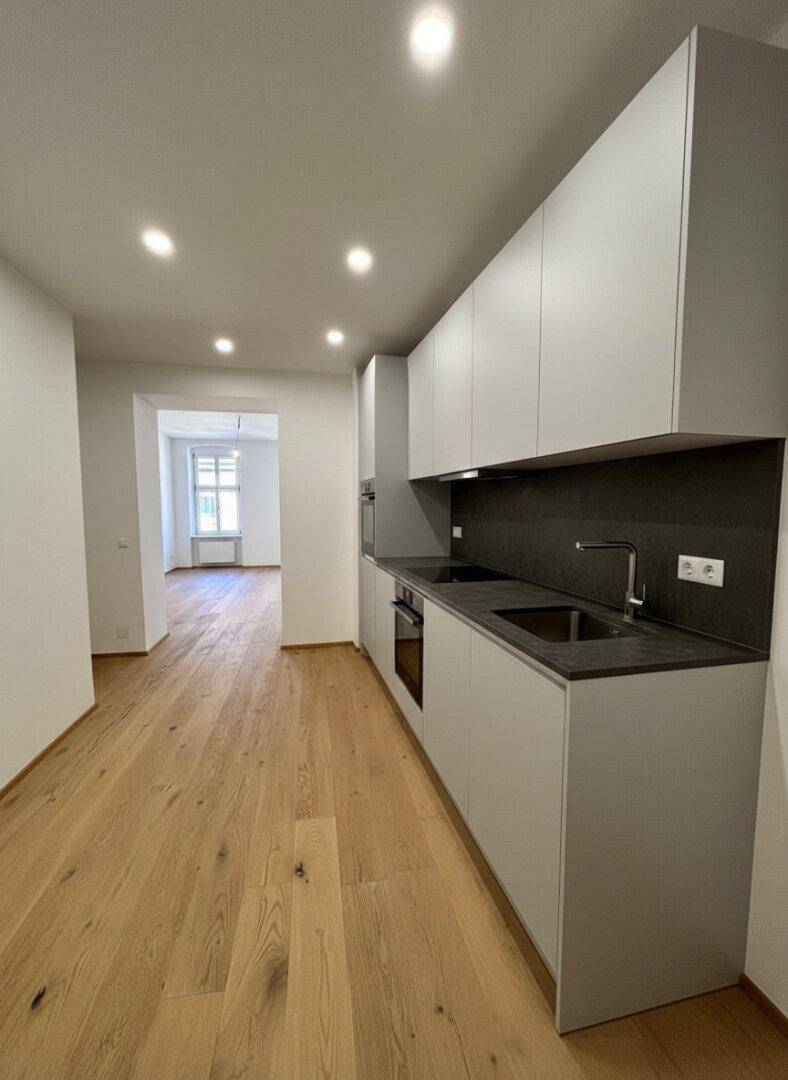 Moderne Einbauküche mit grauen Schränken, dunkler Arbeitsplatte und Holzboden in einem offenen Raum.