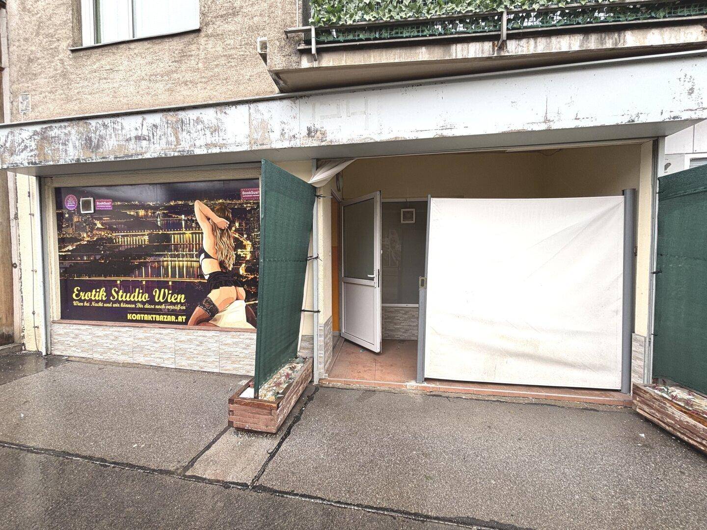 Eingangsbereich eines Erdgeschossgeschäfts mit Werbeplakat und provisorischen Sichtschutzwänden an einer nassen Straße.