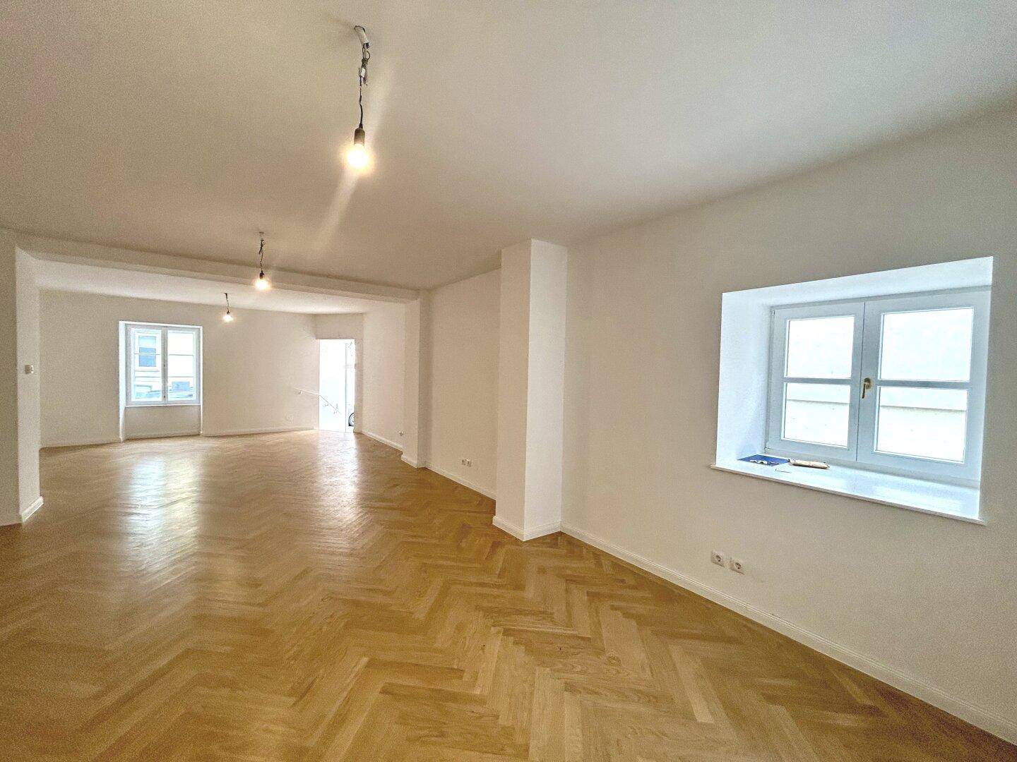 Heller Büroraum mit Fischgrätparkett und zwei Fenstern, die viel Tageslicht hereinlassen, ideal für eine angenehme Arbeitsatmosphäre.