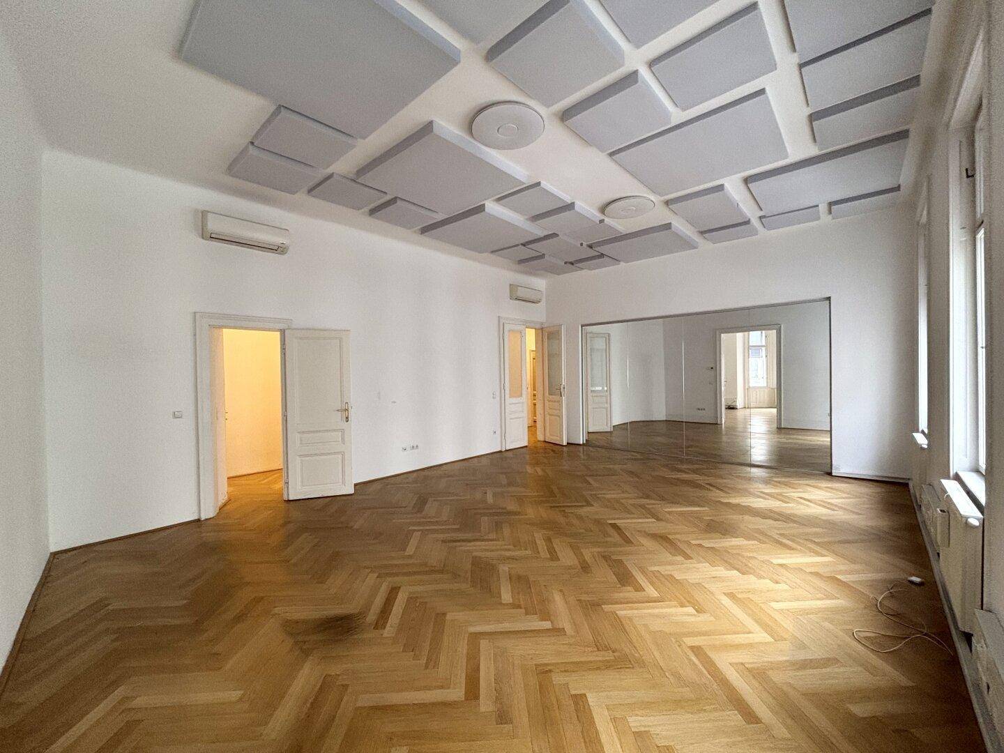 Großer Raum mit Fischgrätparkett, Schalldämmplatten an der Decke und Spiegelflächen.