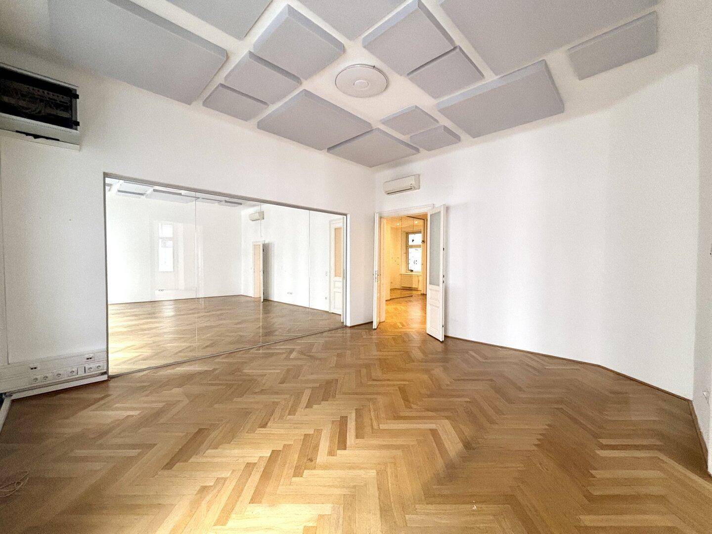 Großer Raum mit Fischgrätparkett, Schalldämmplatten an der Decke und Spiegelflächen.