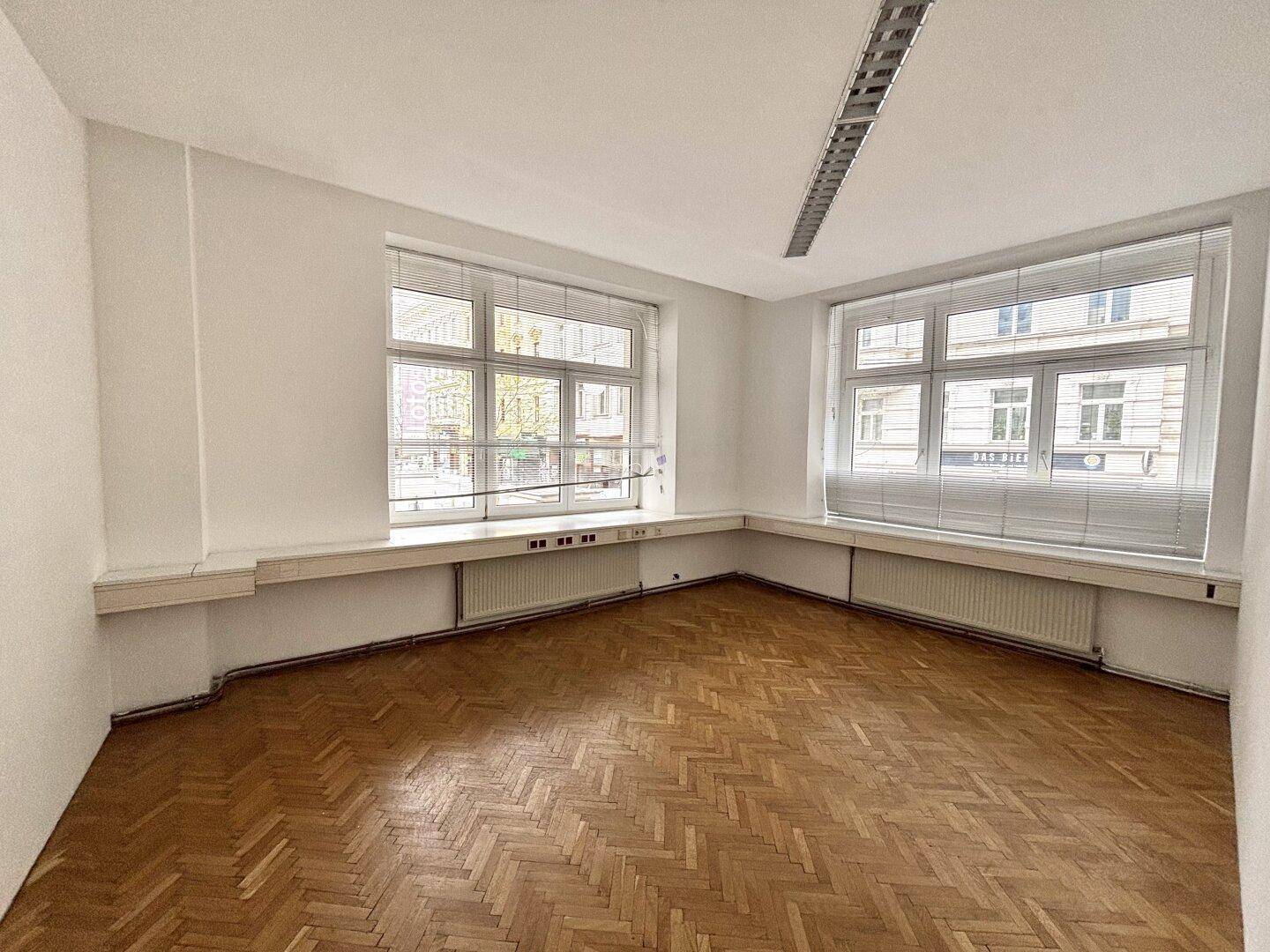 Kleineres Eckbüro mit zwei großen Fenstern, Fischgrätparkett und Heizkörpern unter den Fensterbänken.