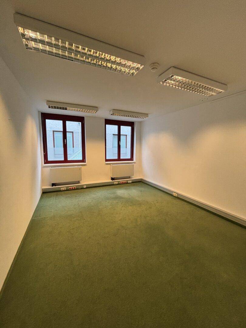 Helles Büro mit grünem Teppichboden, weißen Wänden und zwei Fenstern, ausgestattet mit Heizkörpern.