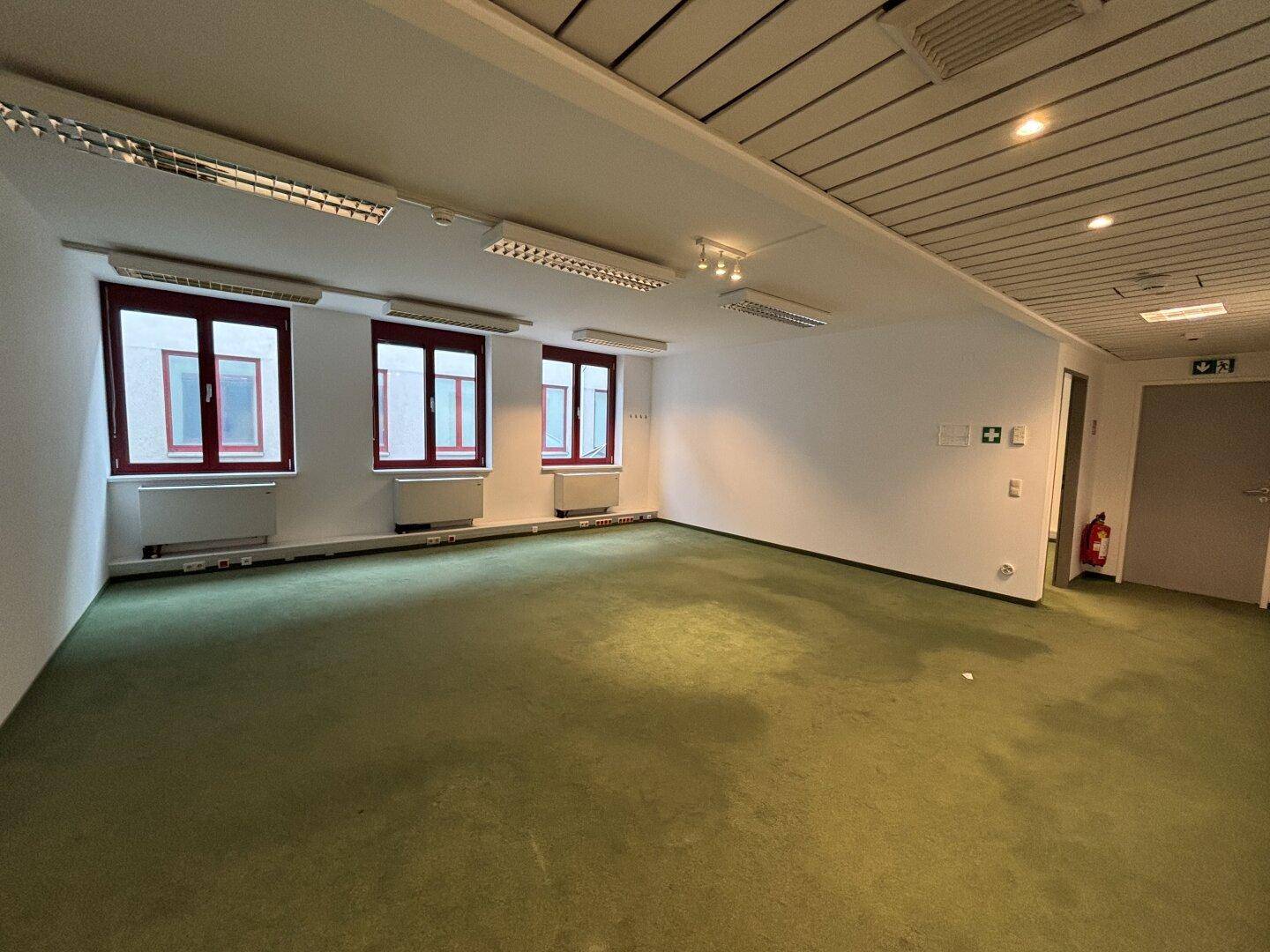 Weitläufiges Büro mit grünem Teppichboden, weißen Wänden und mehreren Fenstern, inklusive Brandschutzvorrichtungen.