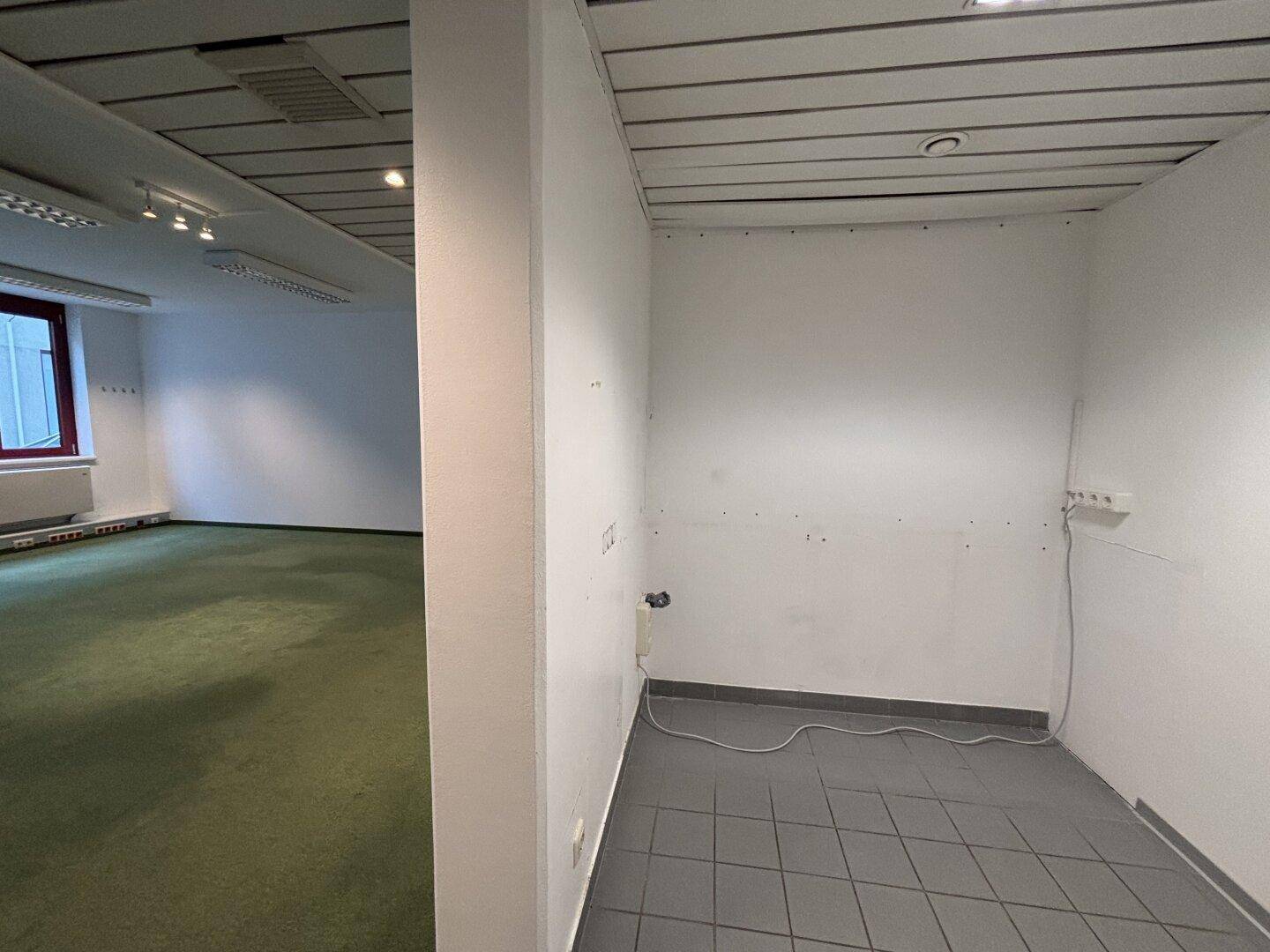 Kleiner Abstellraum mit grauen Fliesen und einer einfachen Ausstattung, angrenzend an einen Bürobereich.