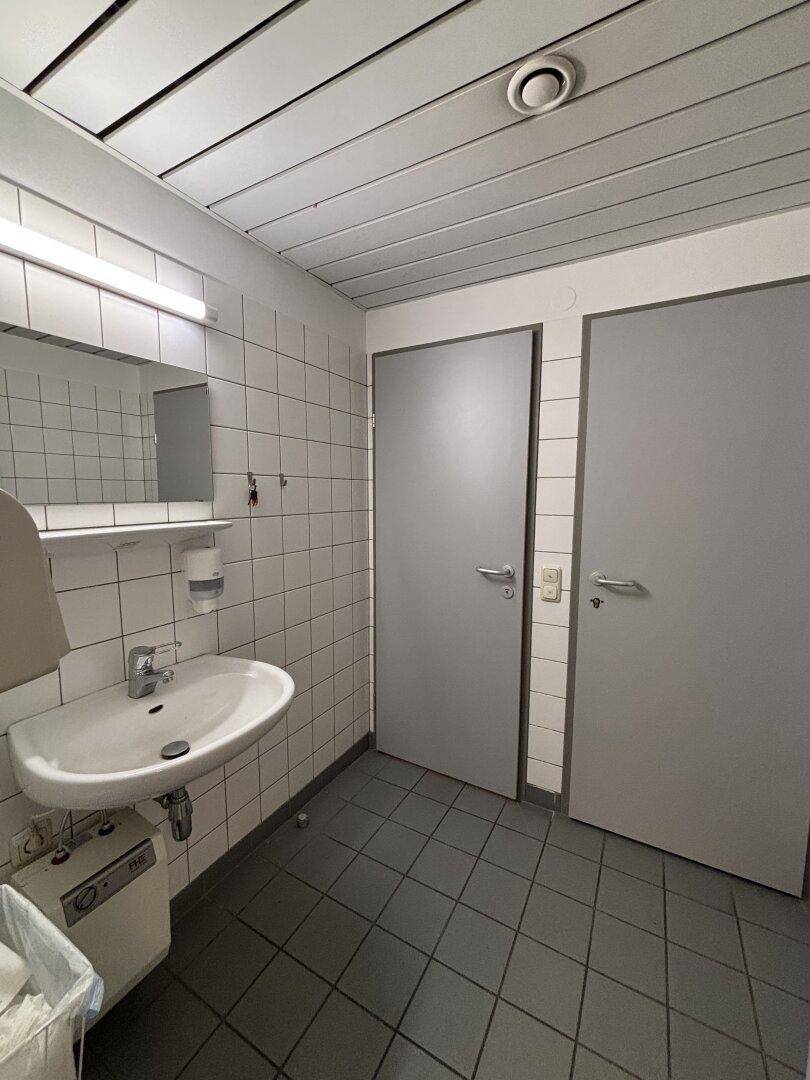 Einfache Toilette mit Waschbecken, Spiegel und weißen Fliesen, ausgestattet mit zwei Türen.