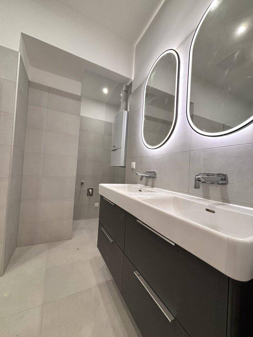 Modernes Badezimmer mit Doppelwaschbecken, beleuchteten Spiegeln und grauen Fliesen.
