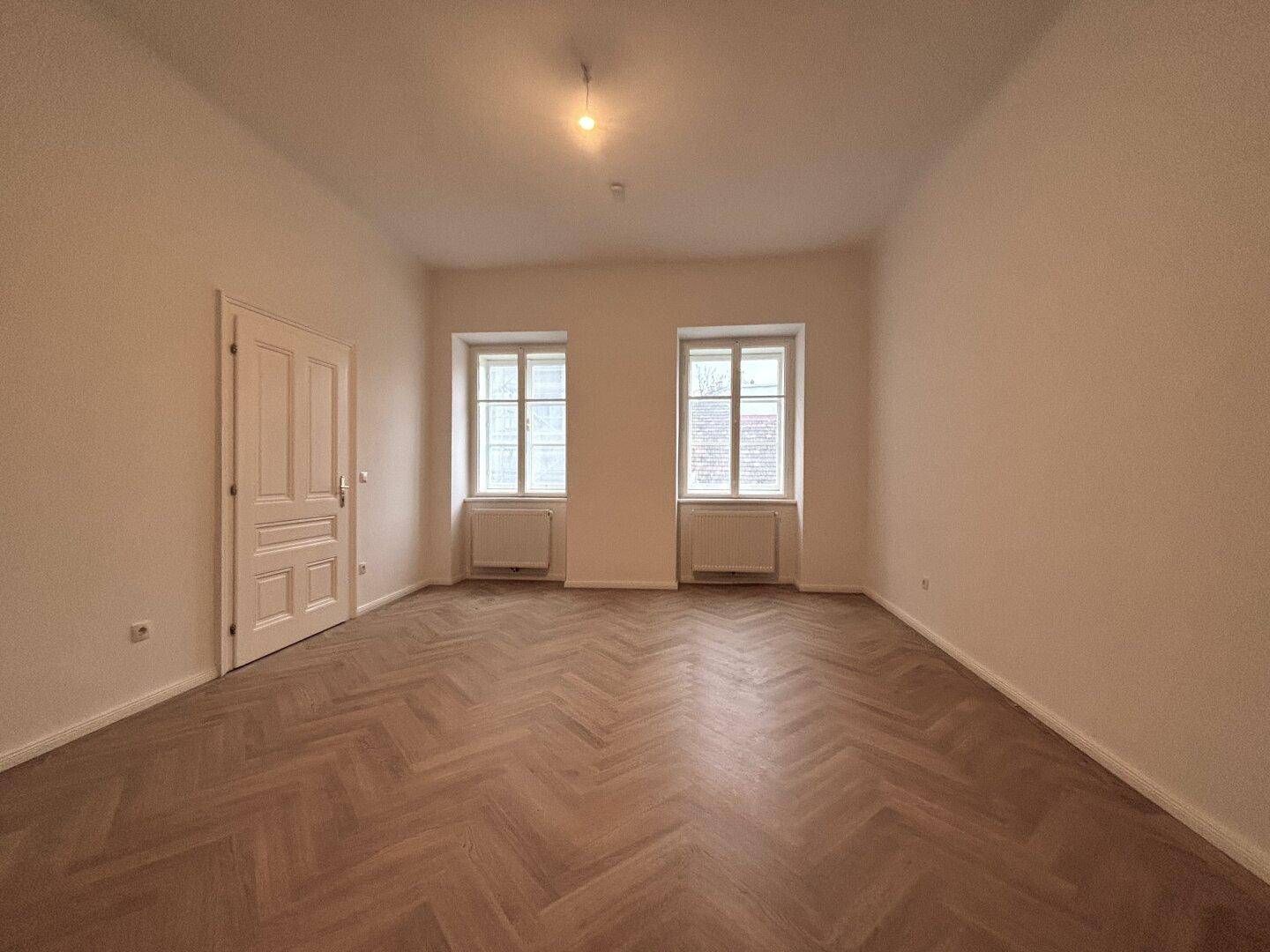 Leerer Raum mit zwei Fenstern, Heizkörpern und Fischgrätparkett.