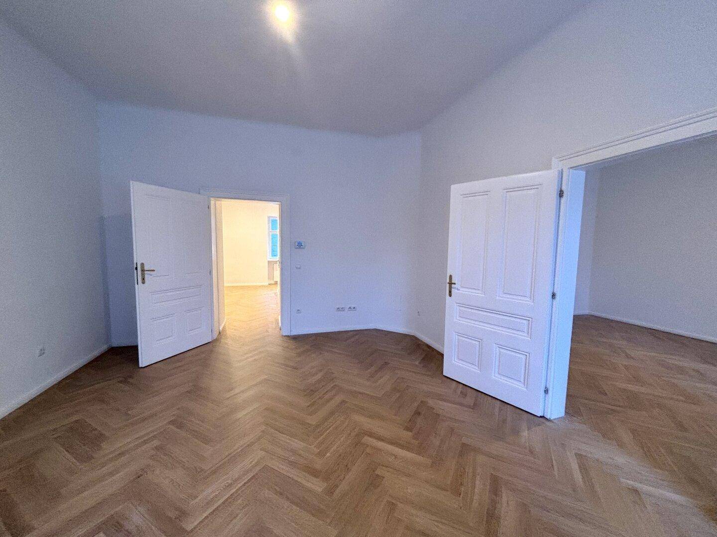 Heller, leerer Raum mit Fischgrätparkett und hohen weißen Türen.