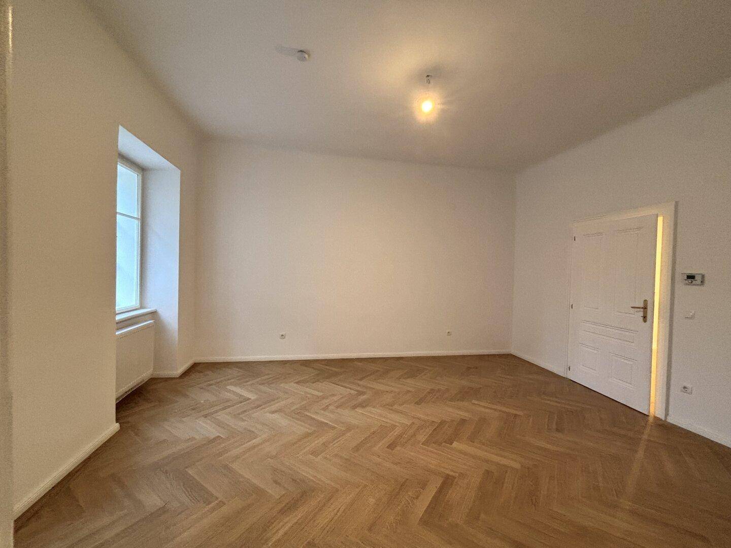 Leerer Raum mit Fischgrätparkett, einem Fenster und weißer Tür.