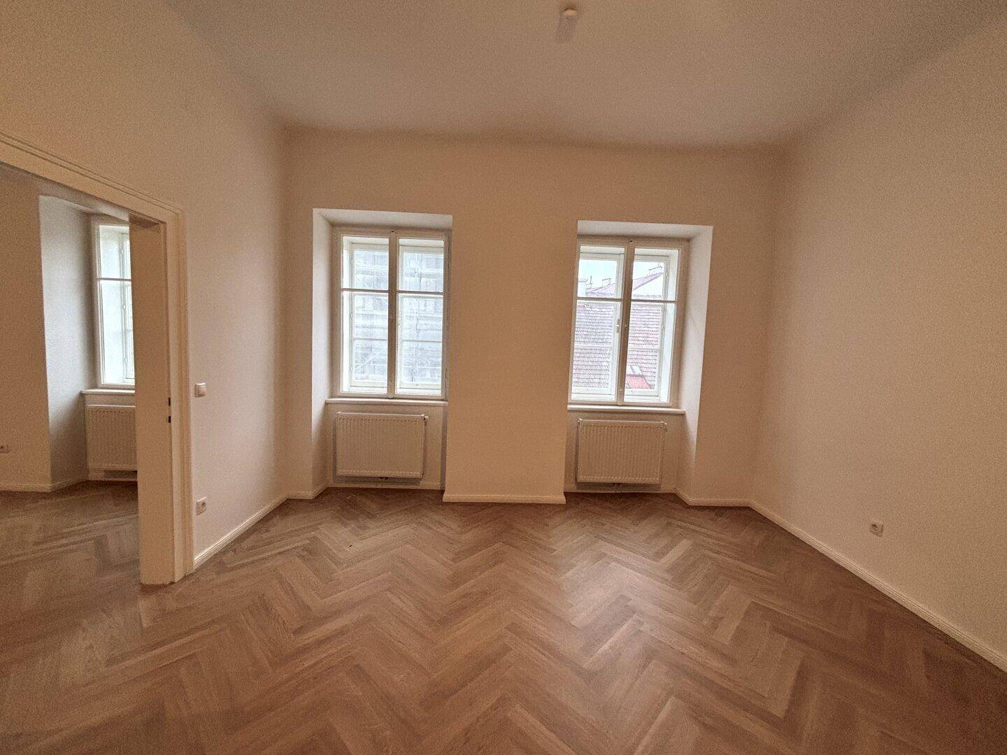 Heller Raum mit Fischgrätparkett, zwei Fenstern und Blick in einen weiteren Raum.