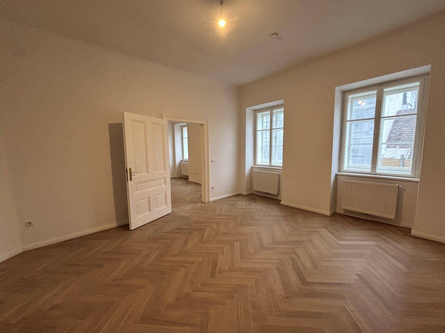 Geräumiger, heller Raum mit Fischgrätparkett, zwei Fenstern und weißen Türen.