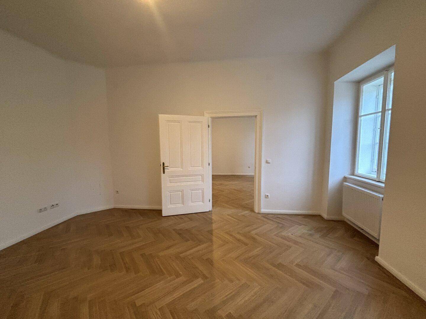 Heller Raum mit Fischgrätparkett, einem Fenster und weißer Tür.