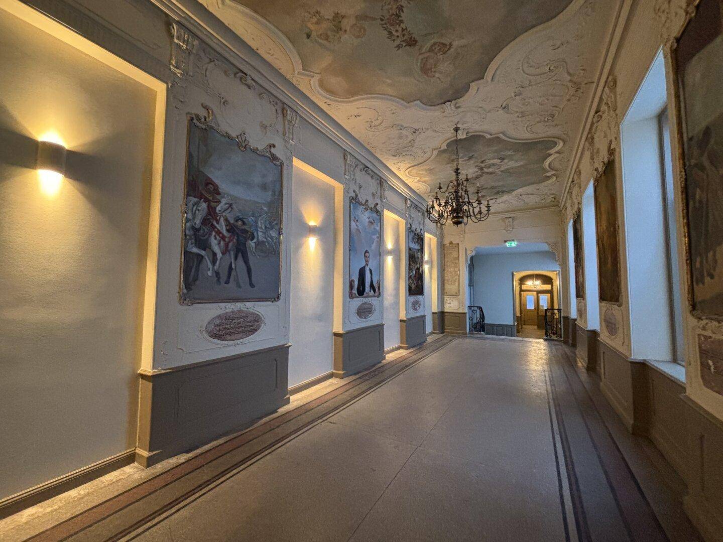 Prunkvoller Flur mit historischen Gemälden, Stuckdecke und elegantem Kronleuchter.
