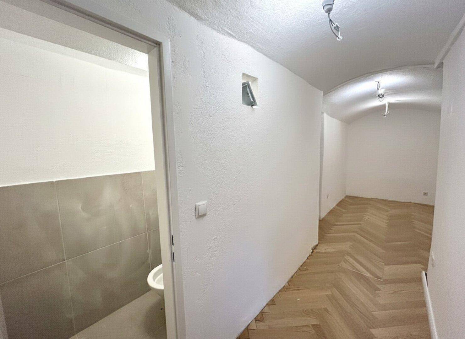 Heller Flur mit Fischgrätparkett, der zu einem kleinen WC-Raum mit Fliesen führt.