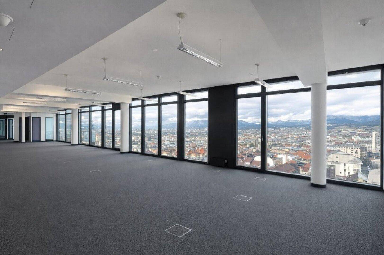 Großer, offener Büroraum mit Teppichboden und raumhohen Fenstern, die einen weiten Stadt- und Bergblick bieten.