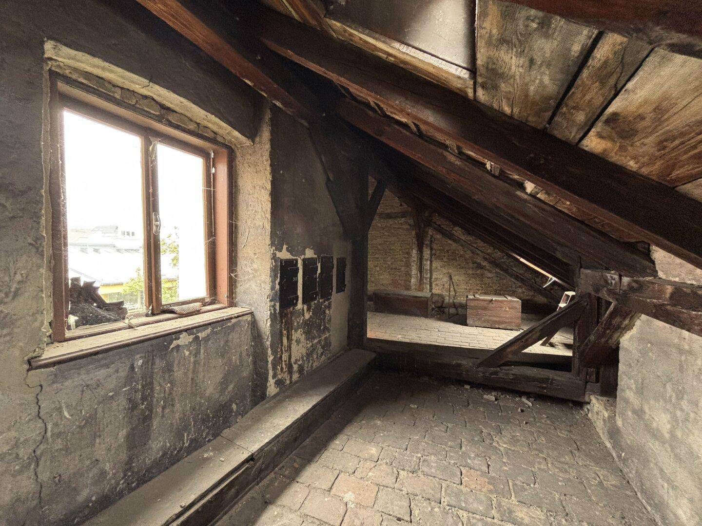 Heller Dachboden mit Ziegelboden, sichtbaren Holzbalken und einem Fenster mit Ausblick.