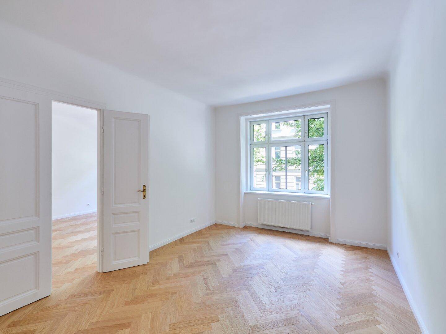 Heller Raum mit Fischgrätparkett, weiß gestrichenen Wänden und einem großen Fenster, das den Raum erhellt.