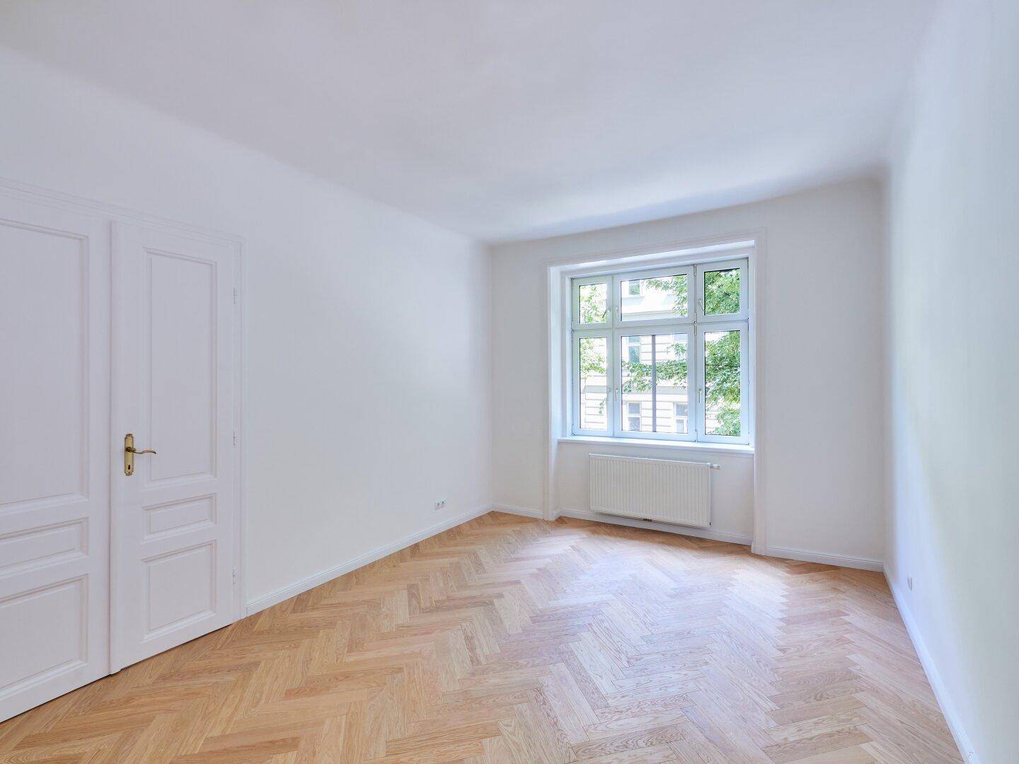 Leerer Raum mit Fischgrätparkett, weiß gestrichenen Wänden und einem großen Fenster, das den Raum erhellt.