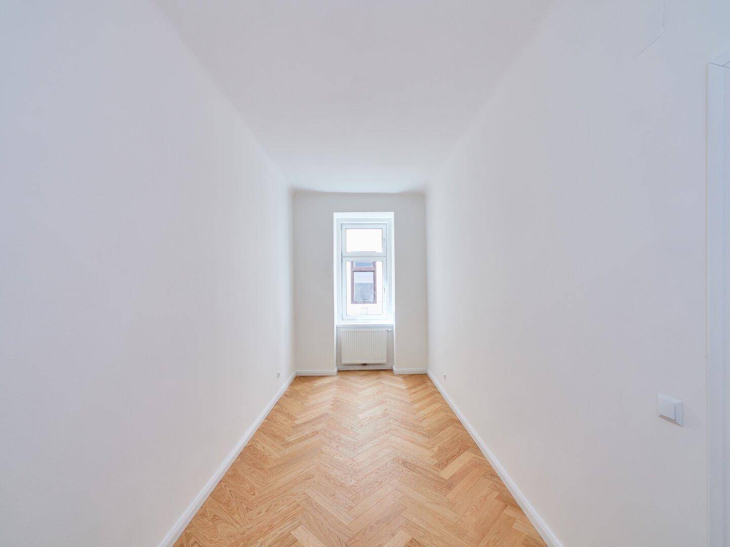 Schmaler Raum mit Fischgrätparkett, weiß gestrichenen Wänden und einem Fenster, ideal als Arbeitszimmer.