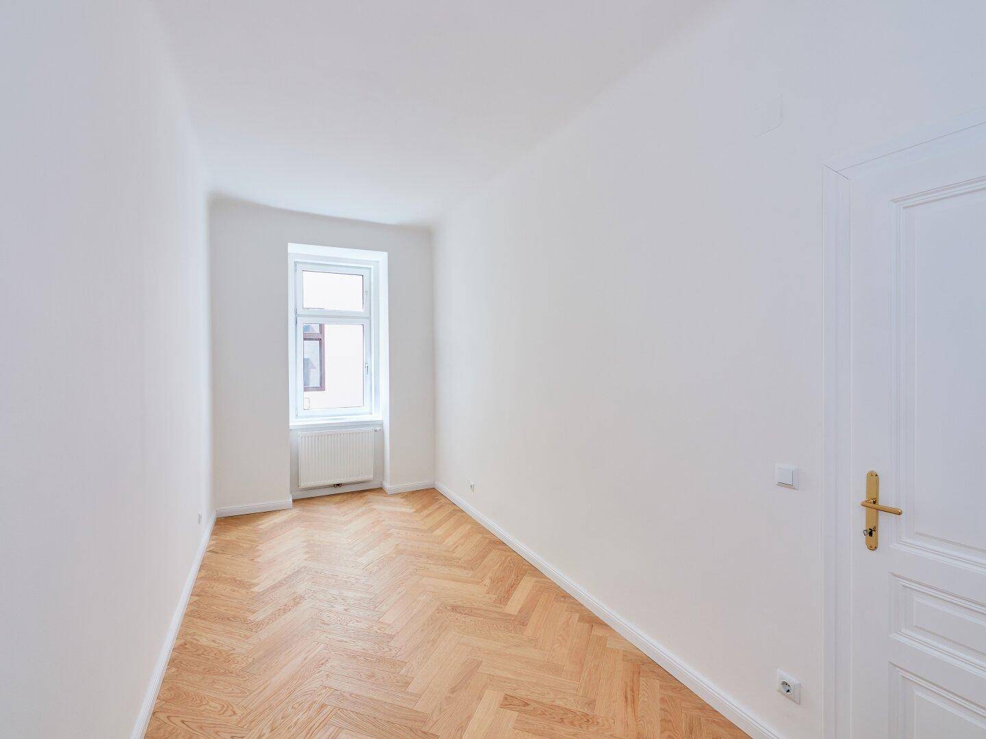 Heller Raum mit Fischgrätparkett, weiß gestrichenen Wänden und einem Fenster, das den Raum erhellt.